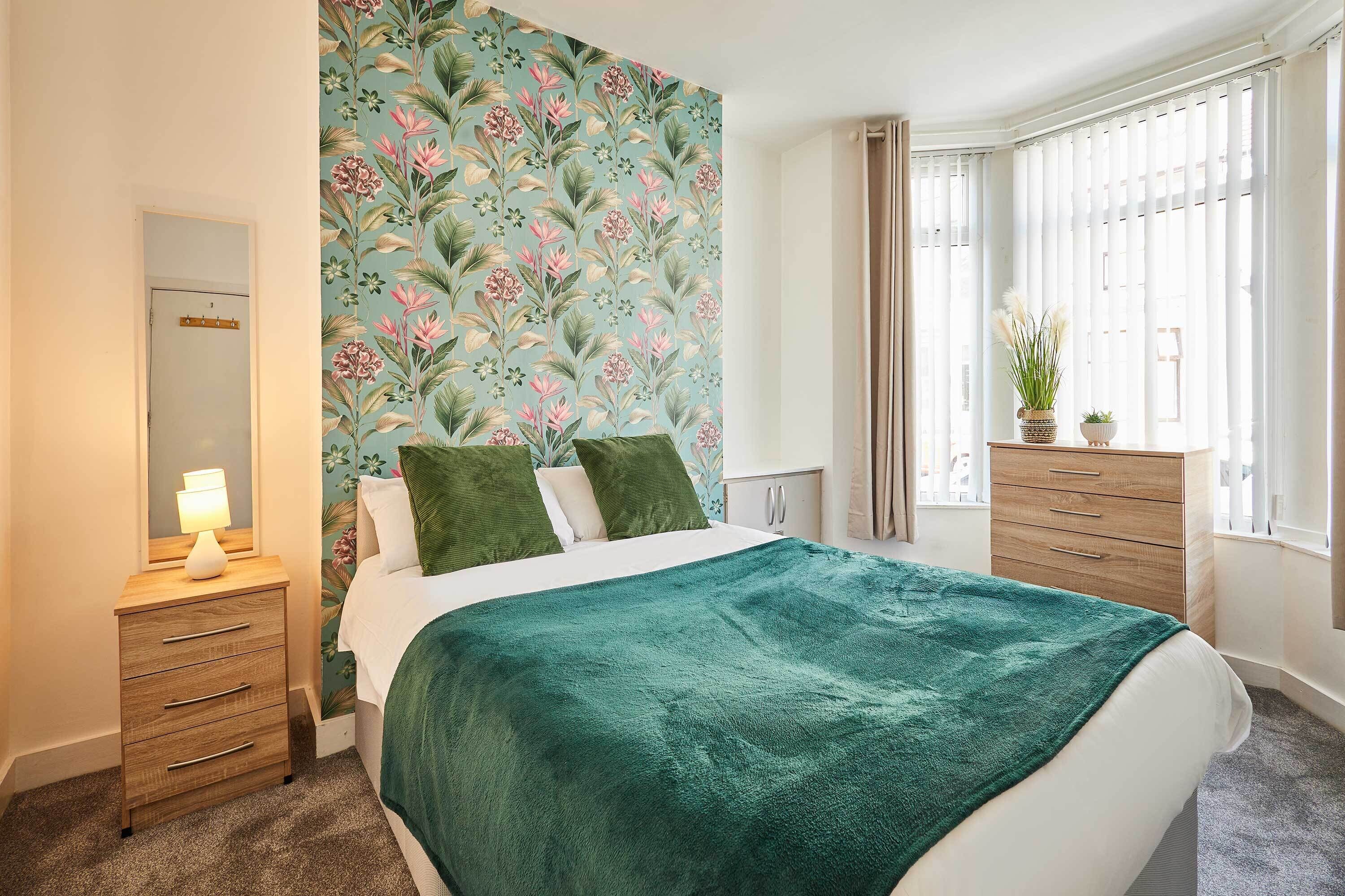A bedroom with a bed, a nightstand, a dresser, a window with curtains, and a floral wallpaper.