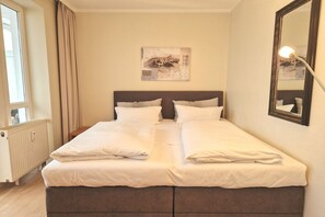 A double bed with white linens, a headboard, a bedside table, a window with curtains, a radiator, a wall-mounted abstract painting, and a mirror with a light. - Komfortable Ferienwohnung fur Zwei in Sellin mit Balkon (Sellin)