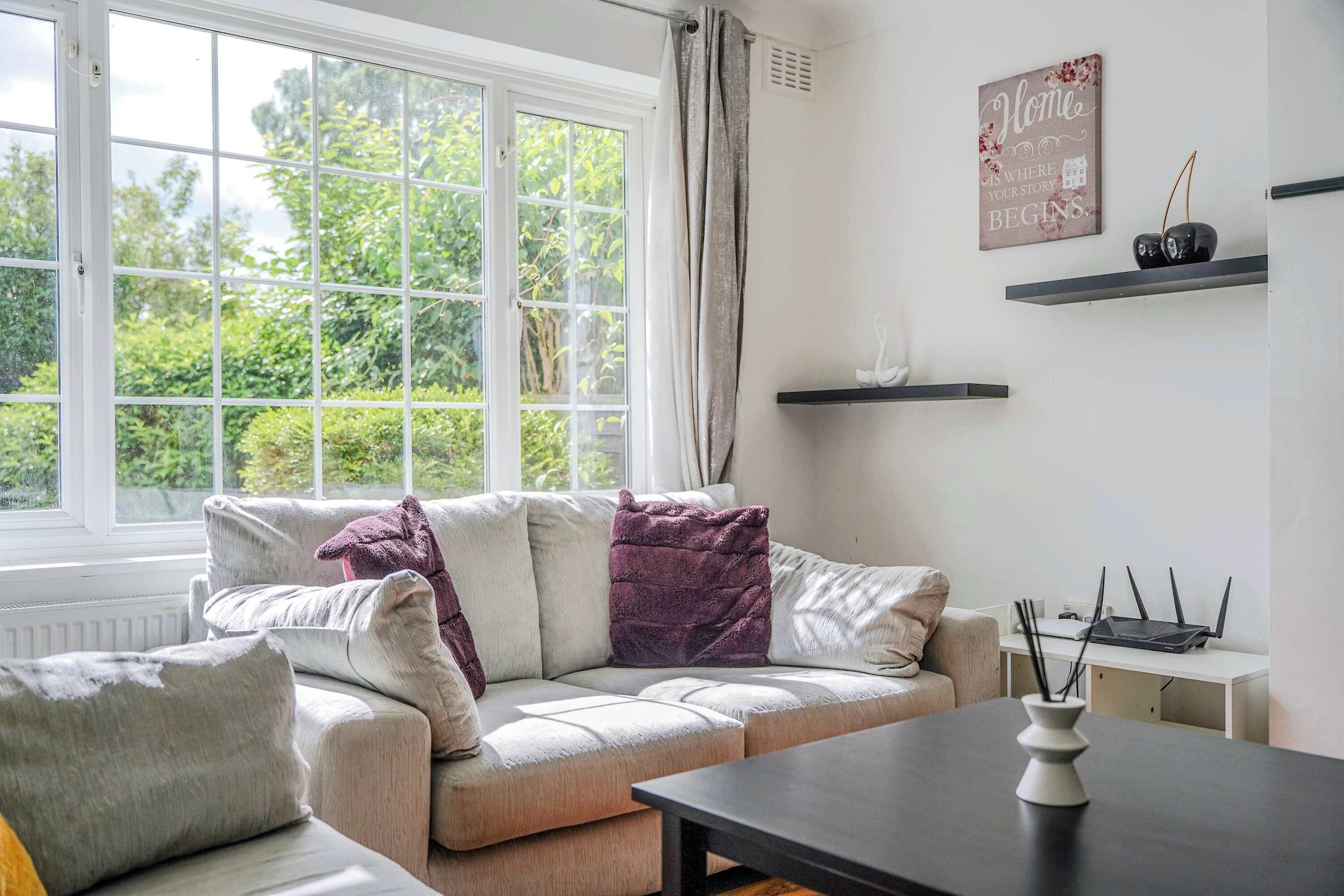 A living room with a sofa, a coffee table, a window with curtains, a wall-mounted shelf with decorative items, and a sign with text.