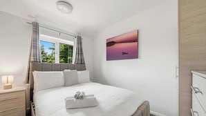 Ein Schlafzimmer mit Bett, Nachttisch, Kommode, Fenster mit Vorhängen und einem Bild an der Wand.