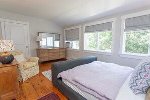 A bedroom with a bed, dresser, chair, and a window with a view of trees. - Vineyard Retreat | Artsy Charm, Deck & Guest Suite (Oak Bluffs)