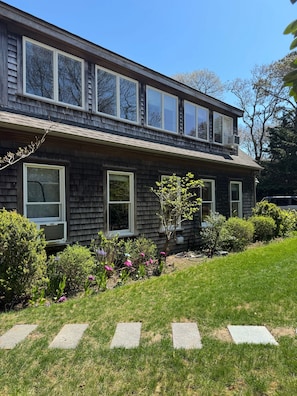 A two-story house with a front yard, a small garden with flowers, and a clear blue sky. - Artsy Vineyard Escape w/ Deck & Guest Suite (Oak Bluffs)
