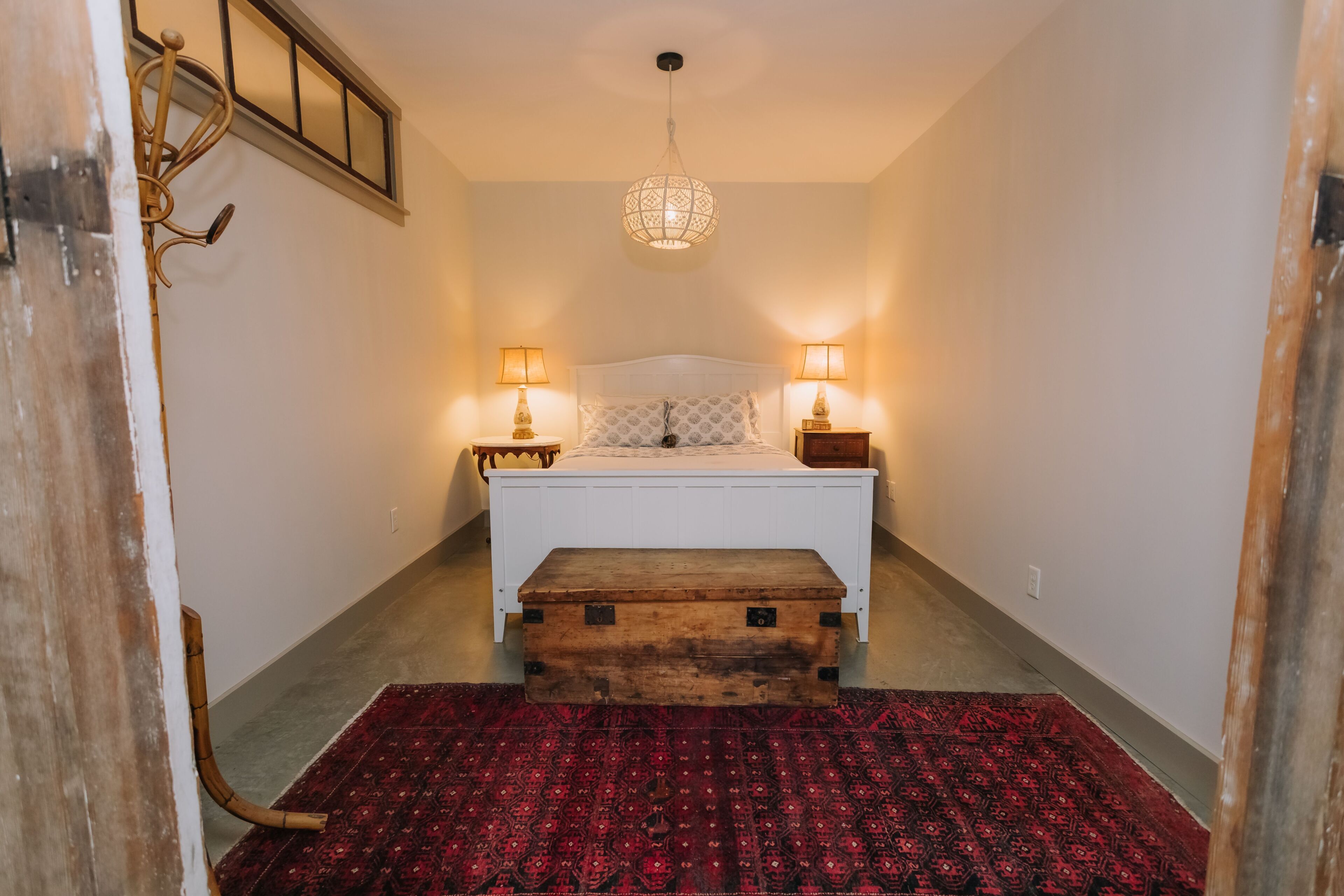 A bedroom with a bed, bedside lamps, a wooden chest, a patterned rug, and a hanging light fixture.