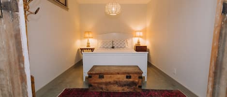 A bedroom with a bed, bedside lamps, a wooden chest, a patterned rug, and a hanging light fixture.