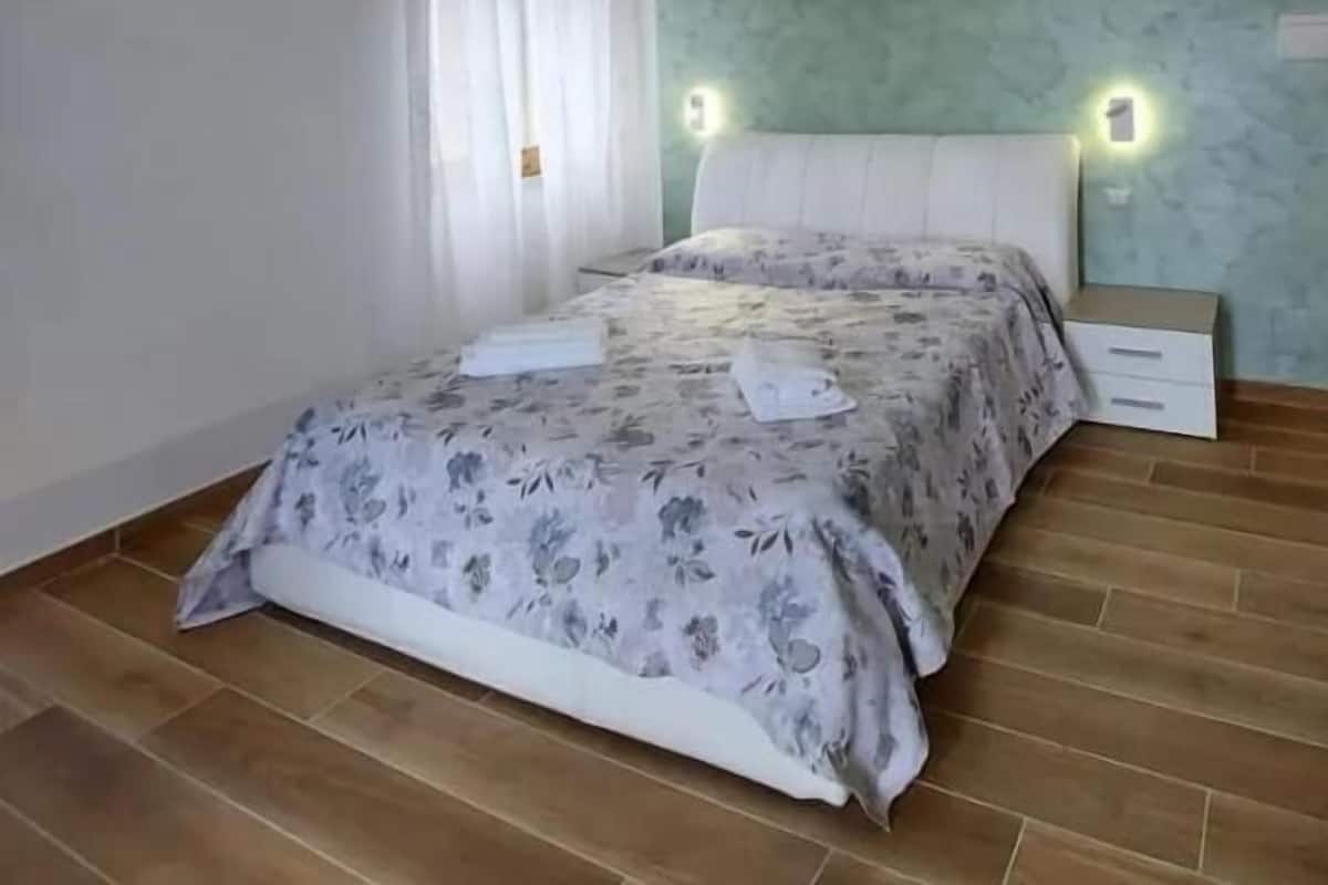 A bedroom with a bed, a nightstand, a wall-mounted light, and a window with curtains.