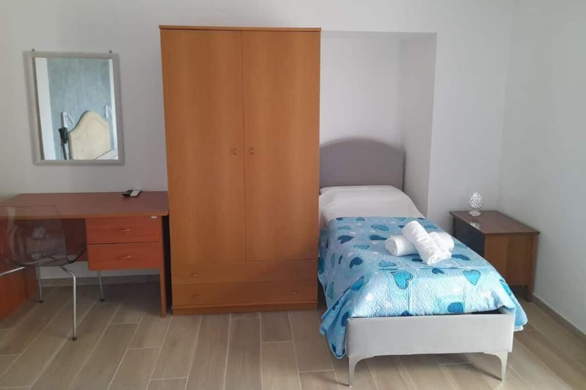 A bedroom with a bed, a wooden wardrobe, a desk, and a chair.