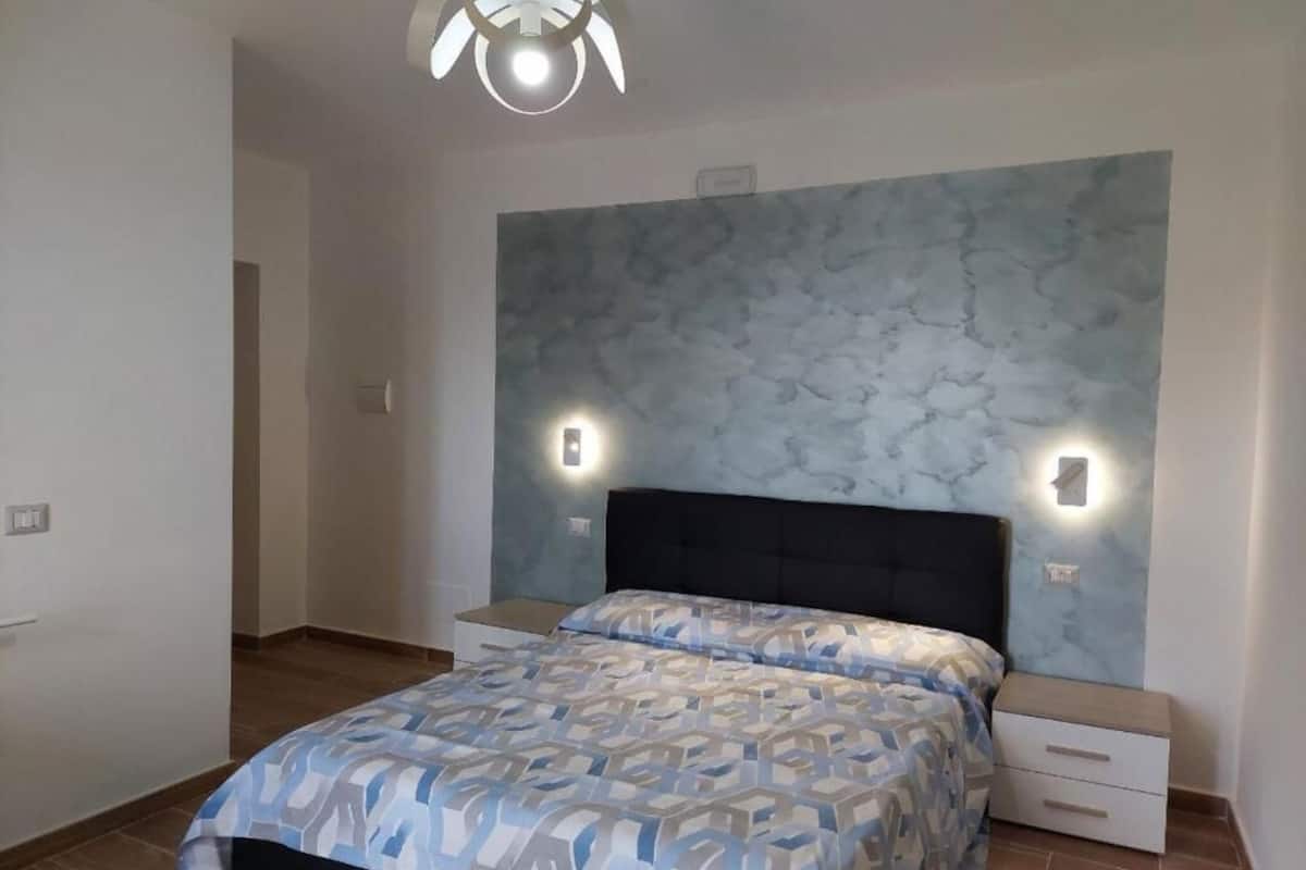 A bedroom with a patterned bedspread, a dark headboard, bedside tables with wall-mounted lights, and a marble-patterned wall.