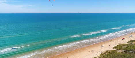 A beach with clear blue water, a sandy shore, and a person paragliding in the distance.