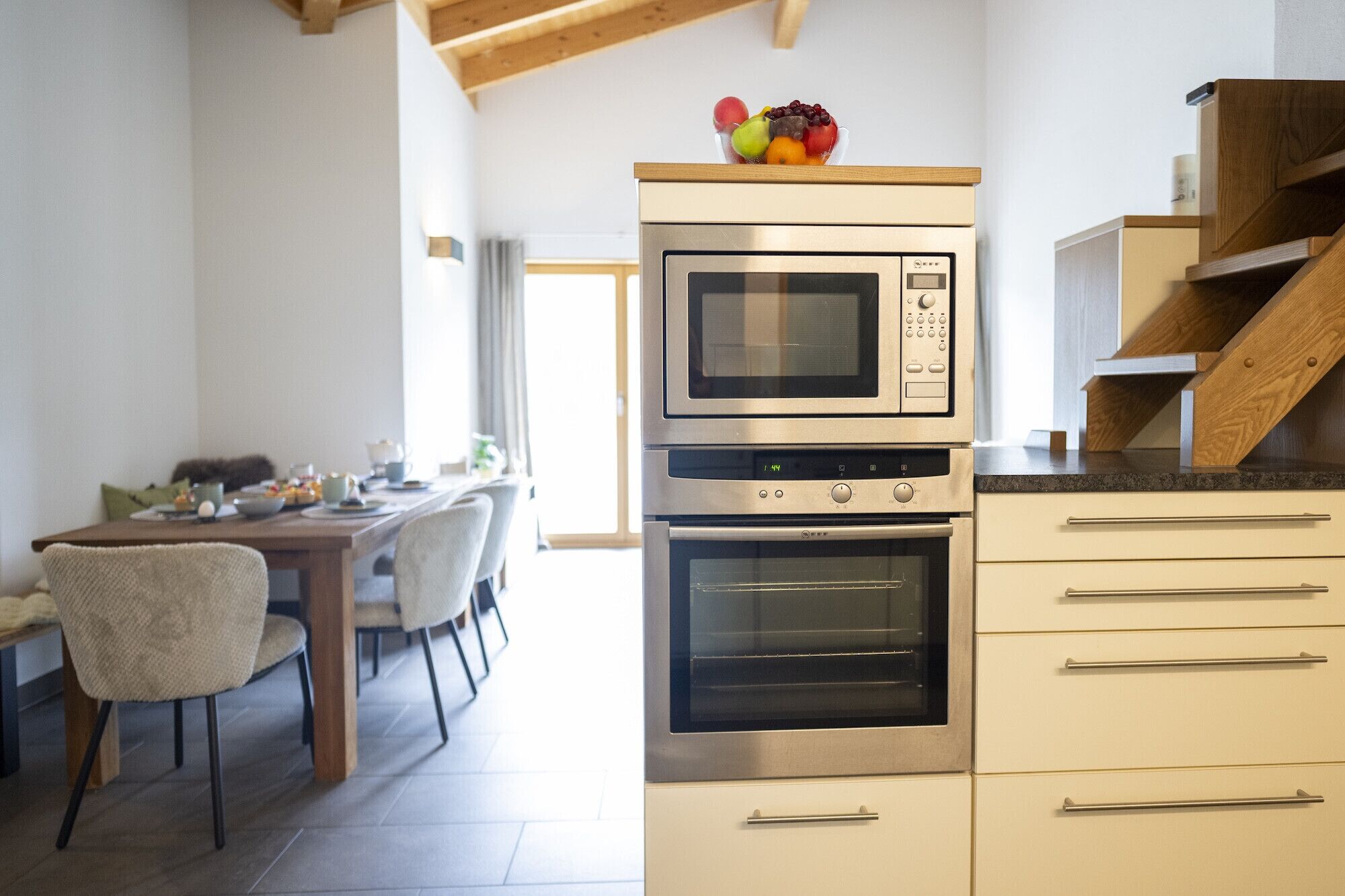 A kitchen with a microwave, oven, and a fruit bowl on the counter. There is a dining table with chairs and a staircase to the right.
