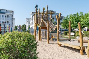 Eine hölzerne Spielkonstruktion mit einer Seilnetzleiter und einer Rutsche, umgeben von Sand und Grünpflanzen.