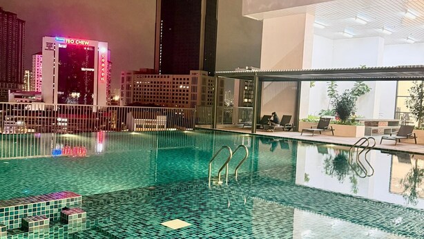 Ein Schwimmbecken mit Fliesenboden, umgeben von einem Zaun, mit Gebäuden im Hintergrund.