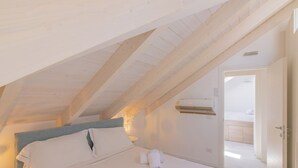 Ein Schlafzimmer mit schrägverdeckter Decke, einem Bett mit weißen Leinentüchern, einer Wand-Klimaanlage und einer Tür zu einem Badezimmer.