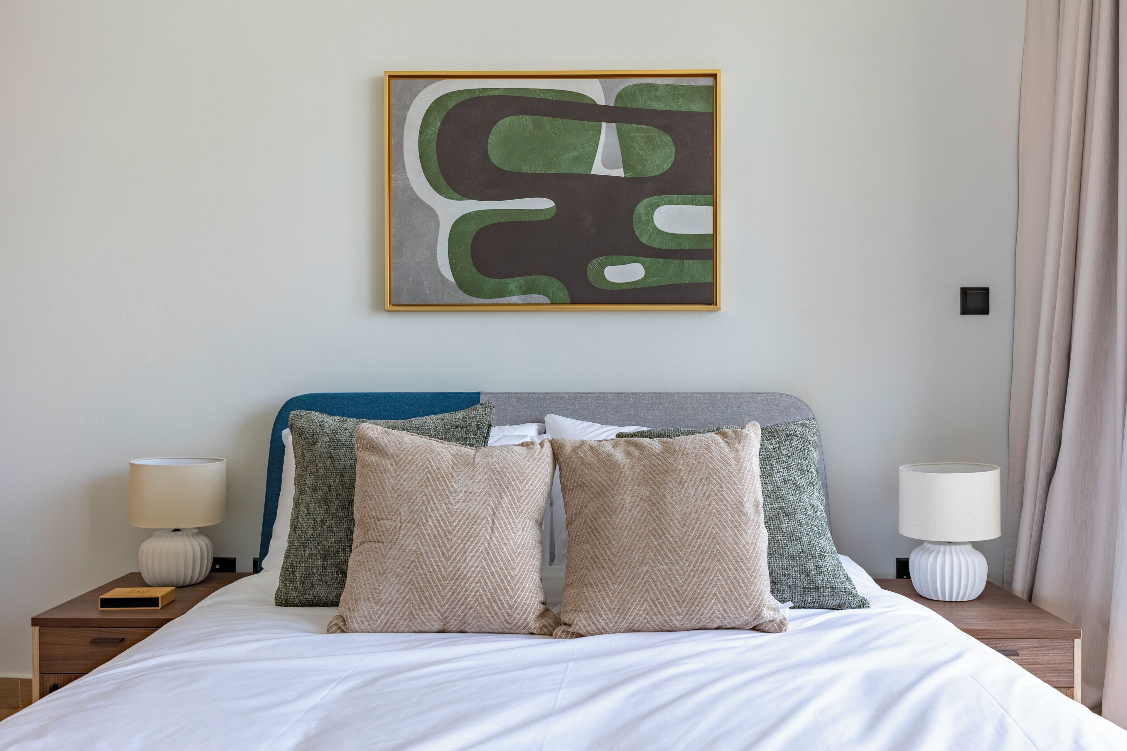A neatly made bed with two pillows, a bedside table with a lamp, and a wall-mounted abstract painting.