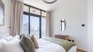 A modern bedroom with a large bed, a nightstand, a mirror, and a view of the city. - Bloomfields 1BR Al Maryah Vista  (Abu Dhabi)