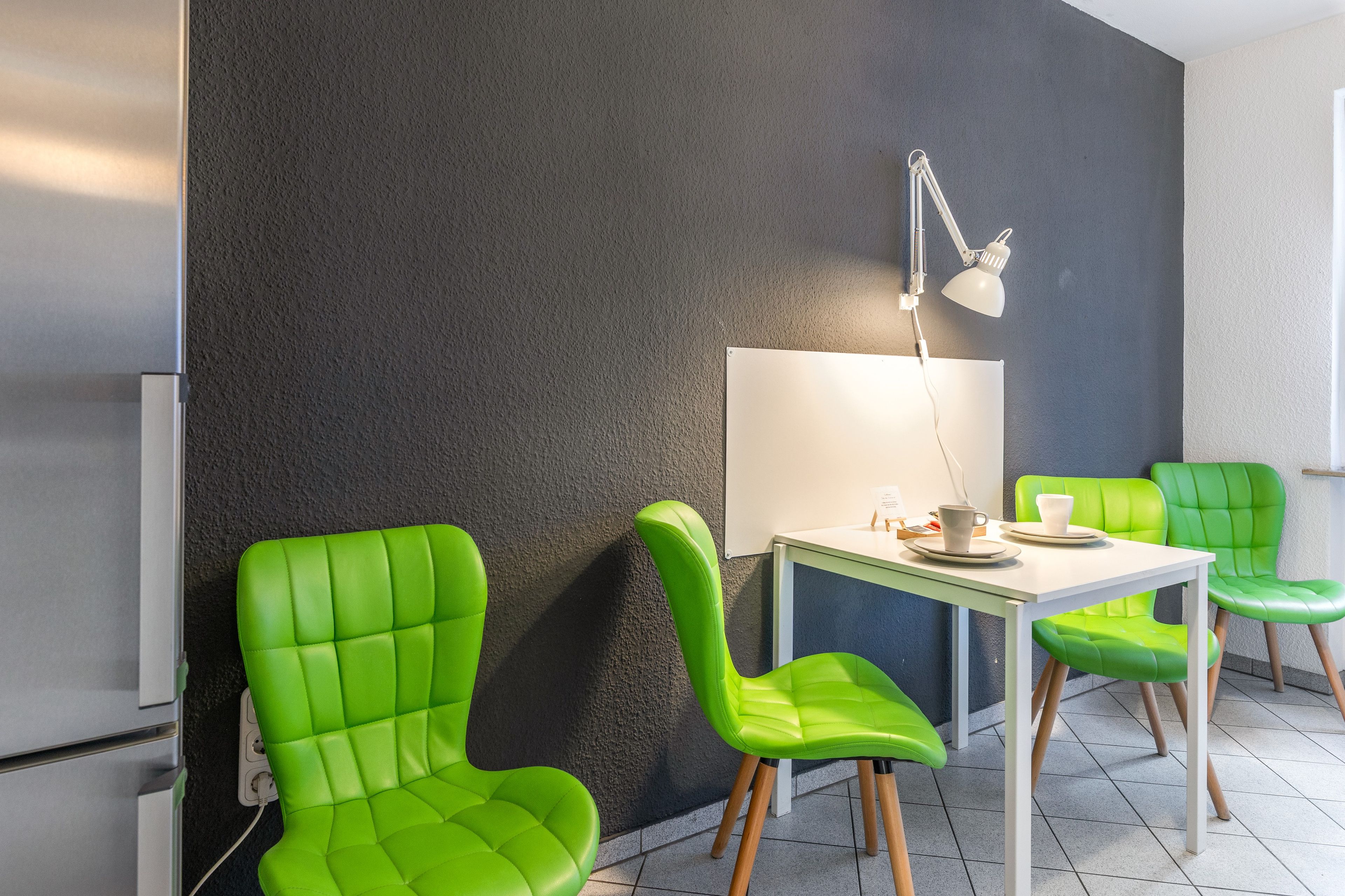A modern room with a white table, two green chairs, a white lamp, and a dark textured wall.