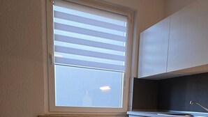 Un comedor con una mesa preparada para dos personas, una ventana con persianas, un radiador y una zona de cocina con armarios.