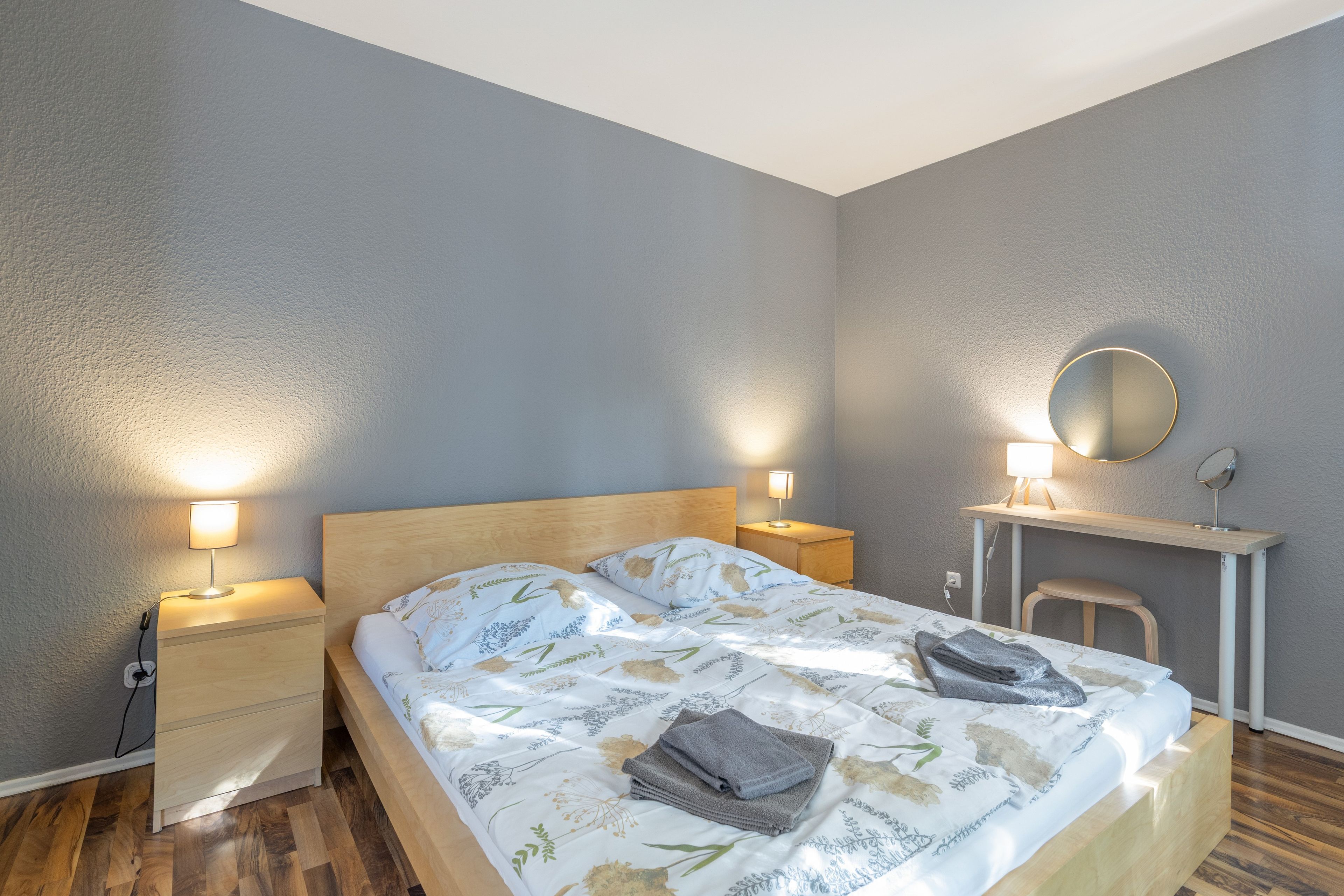 Ein Schlafzimmer mit einem Bett, Nachttischlampen, einem Schreibtisch mit Spiegel und einem Sessel.