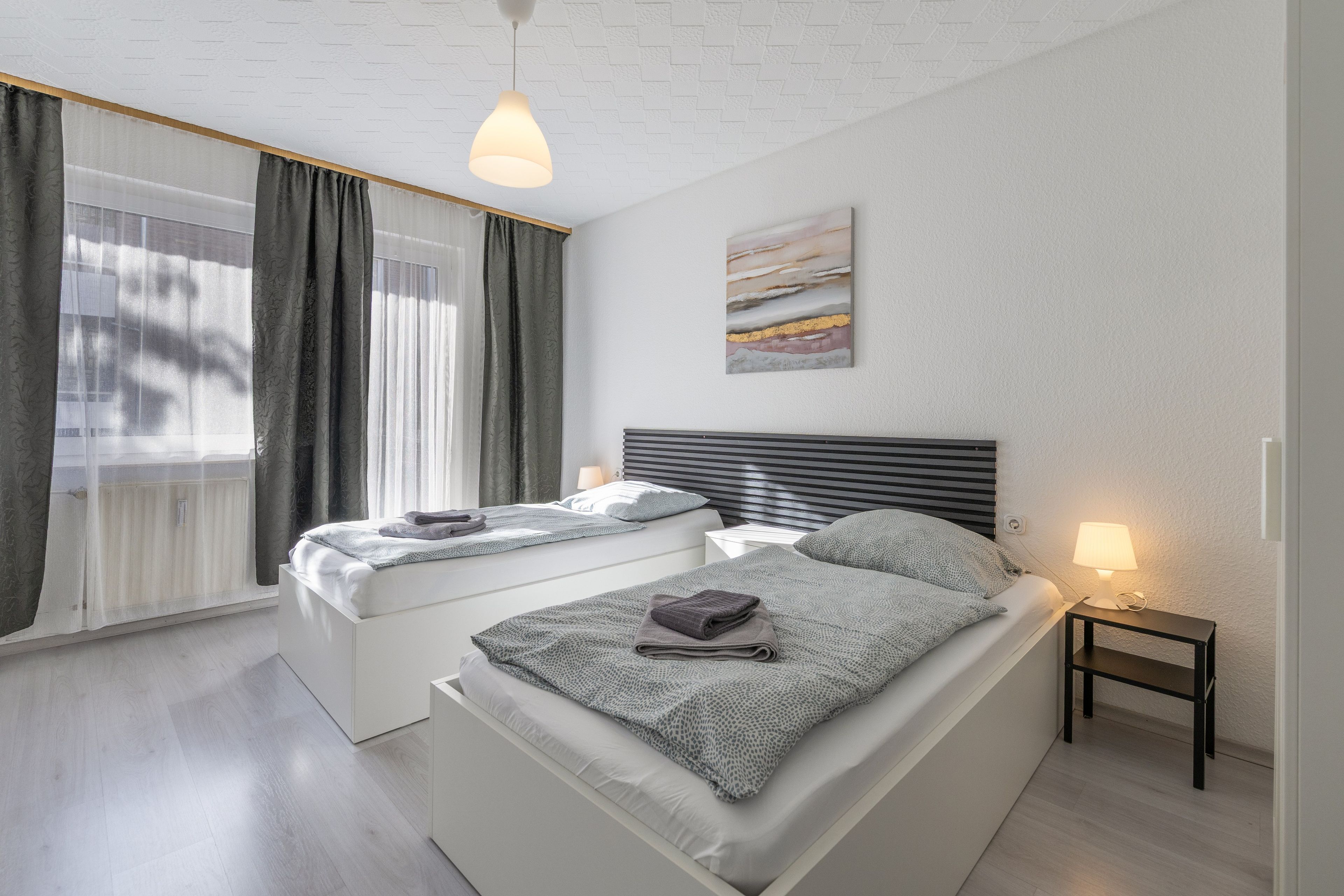 A modern bedroom with two beds, a window with curtains, a painting on the wall, and a bedside lamp.