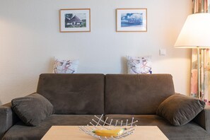 Een bruine bank met twee kussens, een metalen fruitmand op een houten tafel, en twee ingelijste foto's aan de muur.