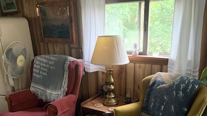 A cozy room with wooden walls, a red armchair, a yellow armchair, a table with a lamp, a fan, and a window with curtains.