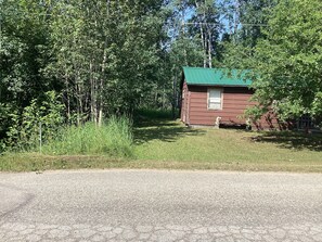 A small, single-story cabin with a green roof, surrounded by dense trees and a gravel road.