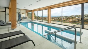 An indoor swimming pool with a view of a coastal landscape, wooden beams, and a sunlit room with exercise equipment. - Pueblo Paraiso Apt, 6 pools, gym & spa (Benahavís)