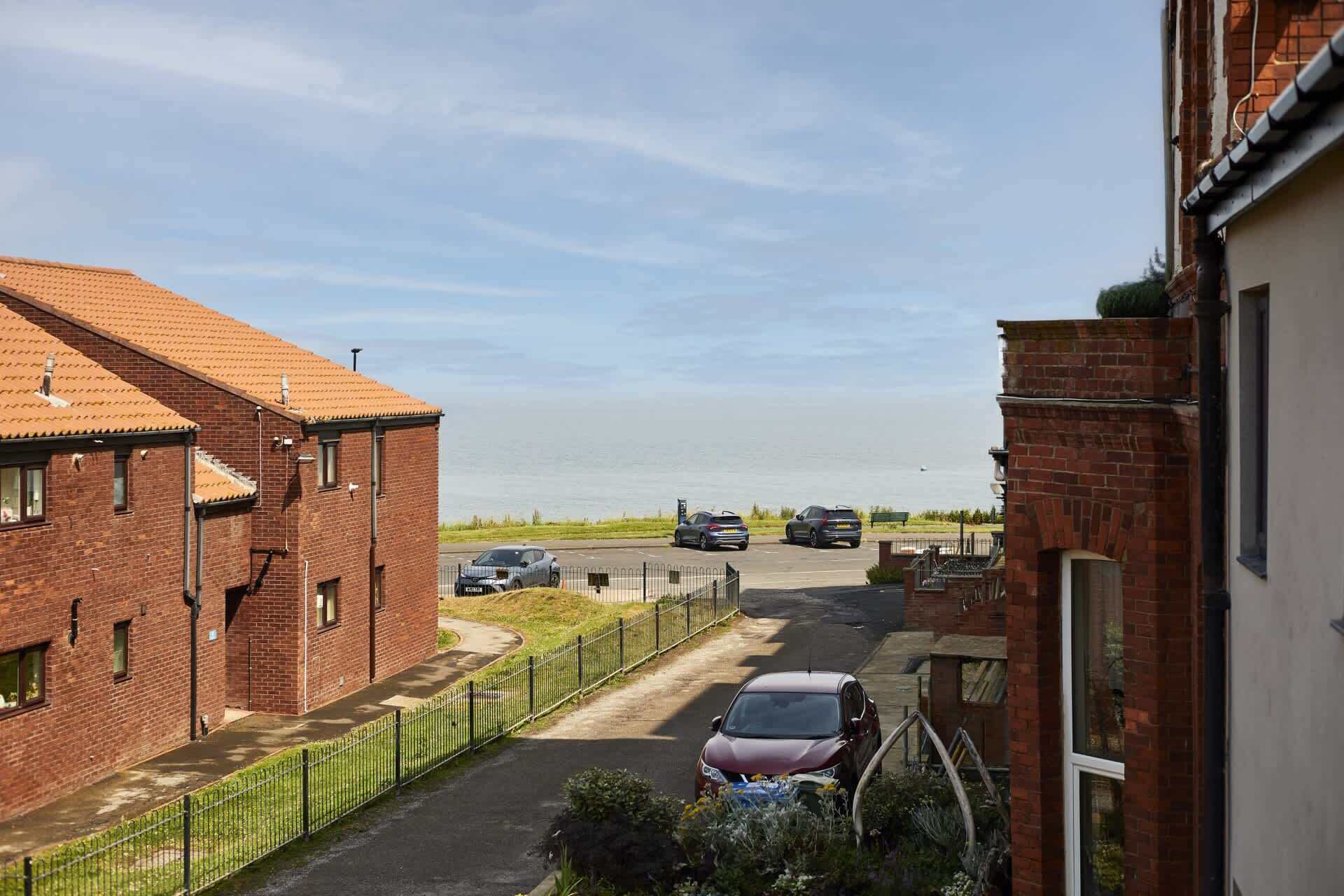 A residential street with brick buildings, a parked car, and a view of the sea.
