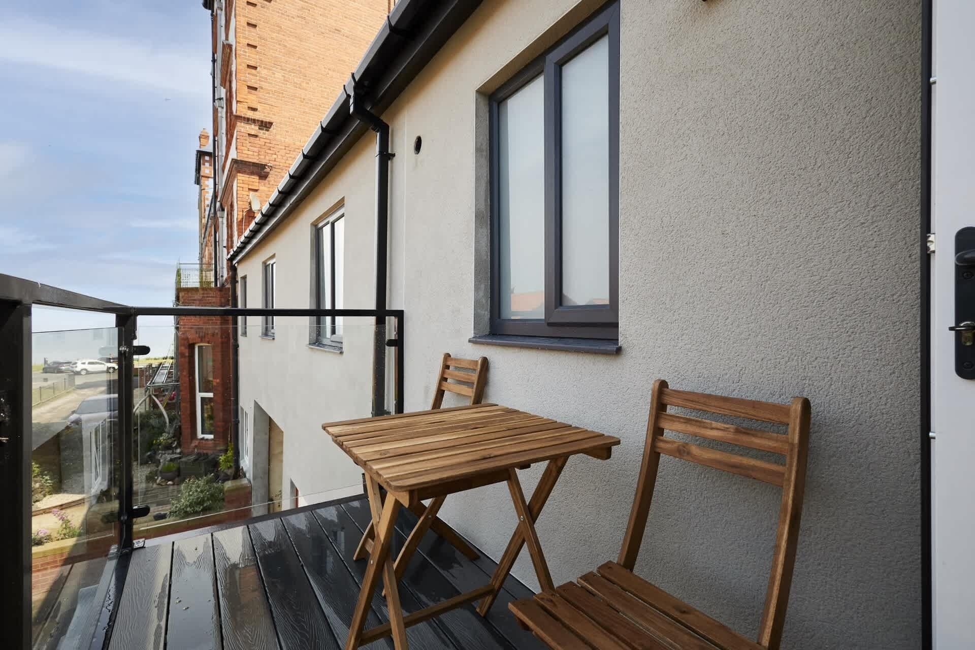 Un balcón con mesa y sillas de madera, una ventana y vista a la calle.