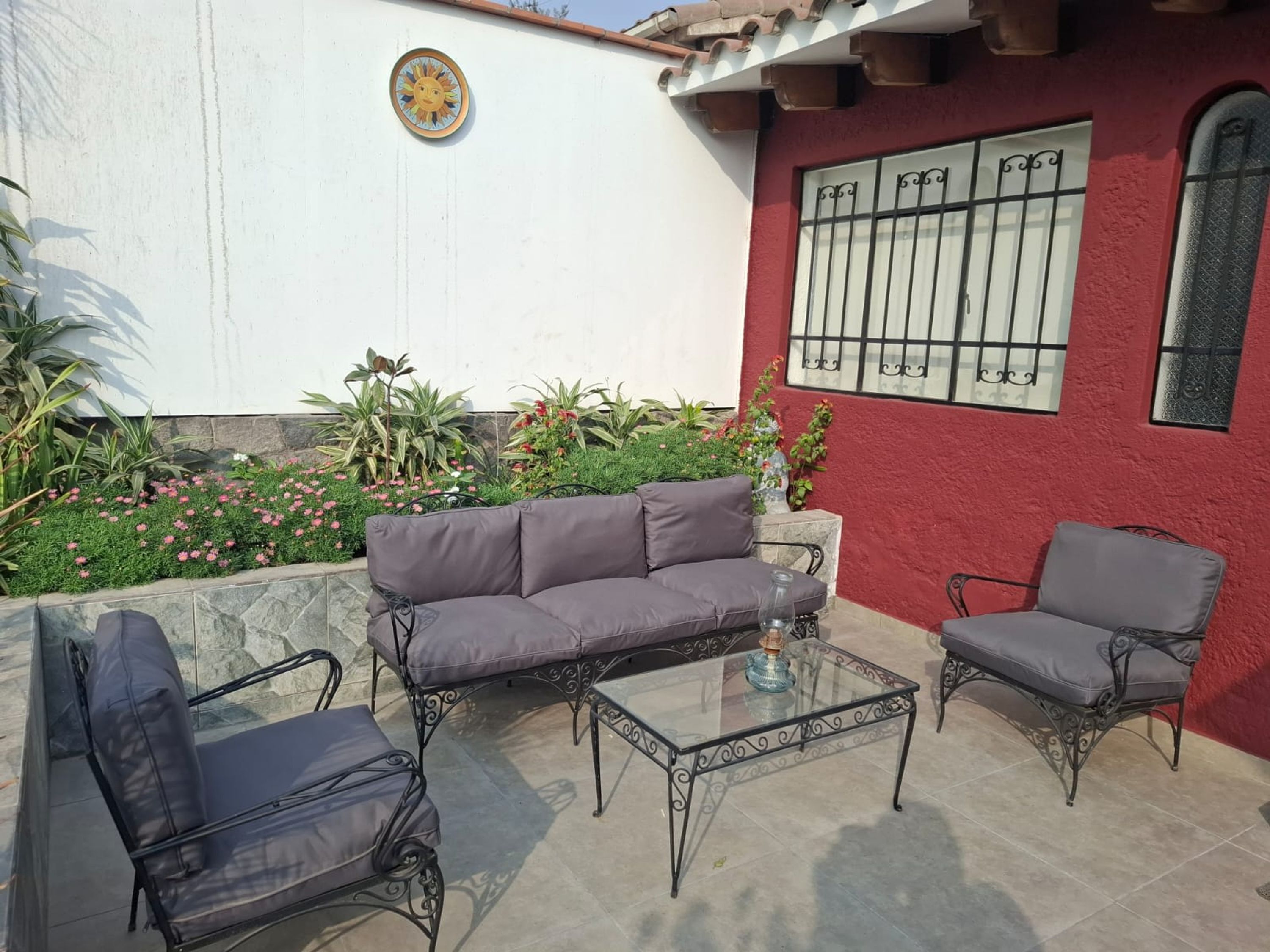 Un patio con un muro rojo, un muro blanco con un adorno de sol, y una ventana con rejas.