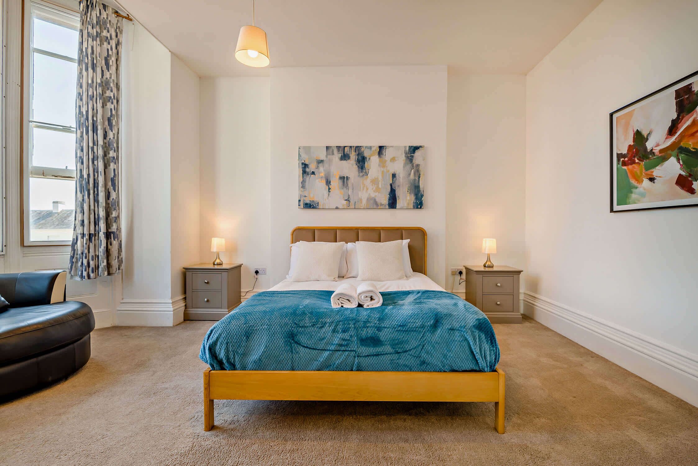 A bedroom with a wooden bed, two bedside tables with lamps, a large abstract painting, and a view of the city through a window with patterned curtains.