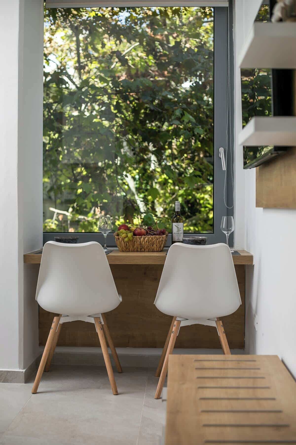 A modern kitchen with a large window, two white chairs, a wooden table, and a fruit basket with wine glasses.