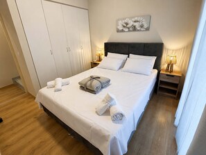 A neatly made bed with white linens, a grey pillow, and rolled towels. There are two bedside tables with lamps, a grey headboard, and a white wardrobe. - Beachside Hideaway - Steps From Lechaio Blue Shore (Corinth)
