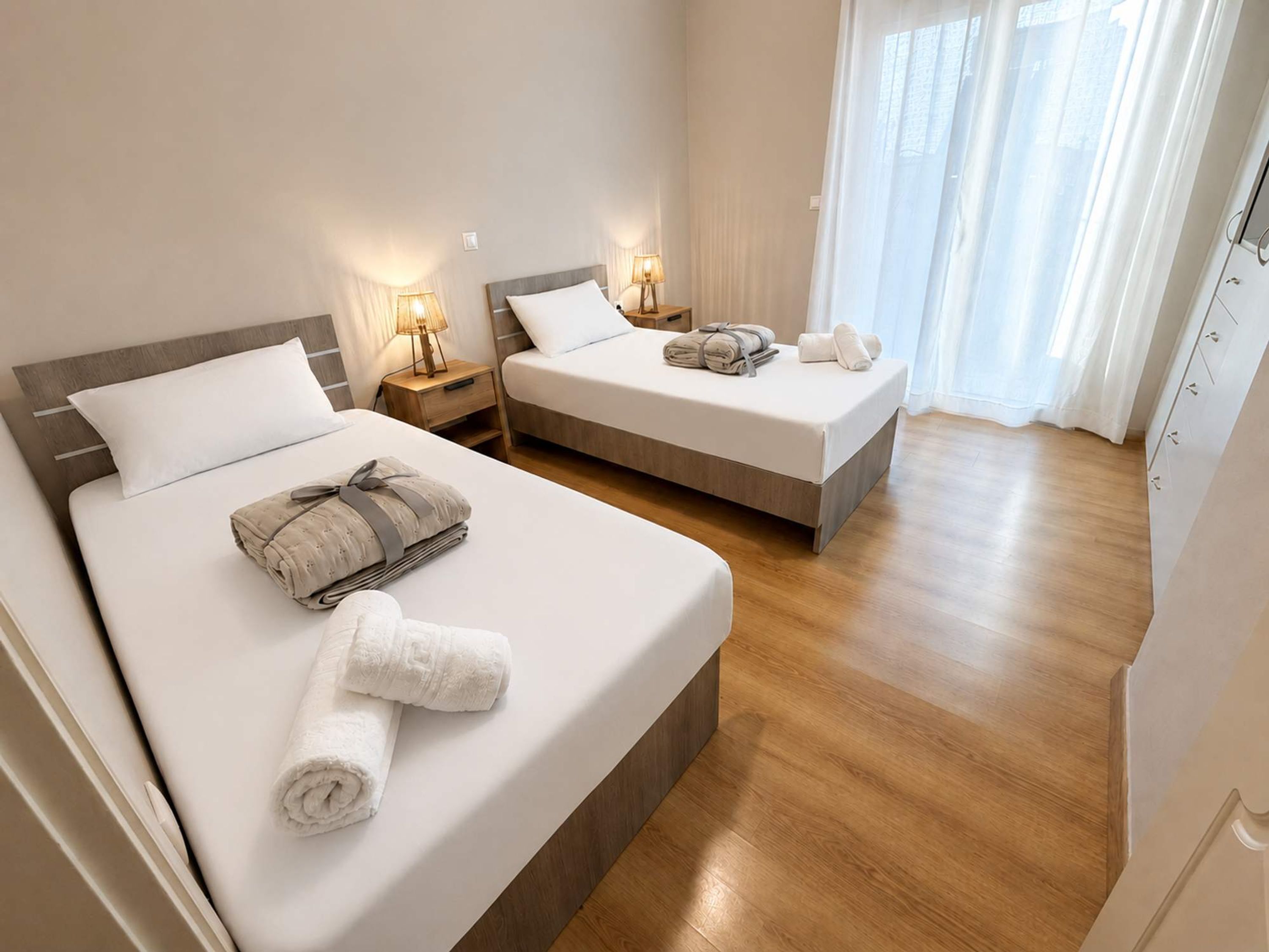 A hotel room with two beds, bedside lamps, a wooden floor, and a window with curtains.