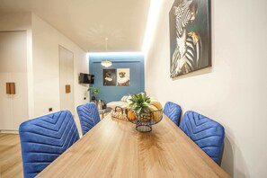 A dining area with a wooden table, blue upholstered chairs, a plant centerpiece, and artwork on the walls. - Noble Heraklion Suites Crete Azure Suite (Heraklion)