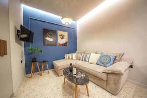 A living room with a blue accent wall, a sofa with patterned cushions, a small black table with a vase and glasses, and a mounted TV. - Noble Heraklion Suites Crete Azure Suite (Heraklion)