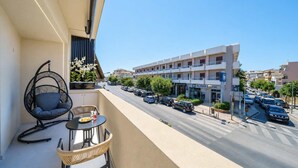 A balcony with a hanging chair, a small table with a plate and glasses, and a view of a street with parked cars and a building with signage. - Noble Heraklion Suites Terra Suite in Crete (Heraklion)