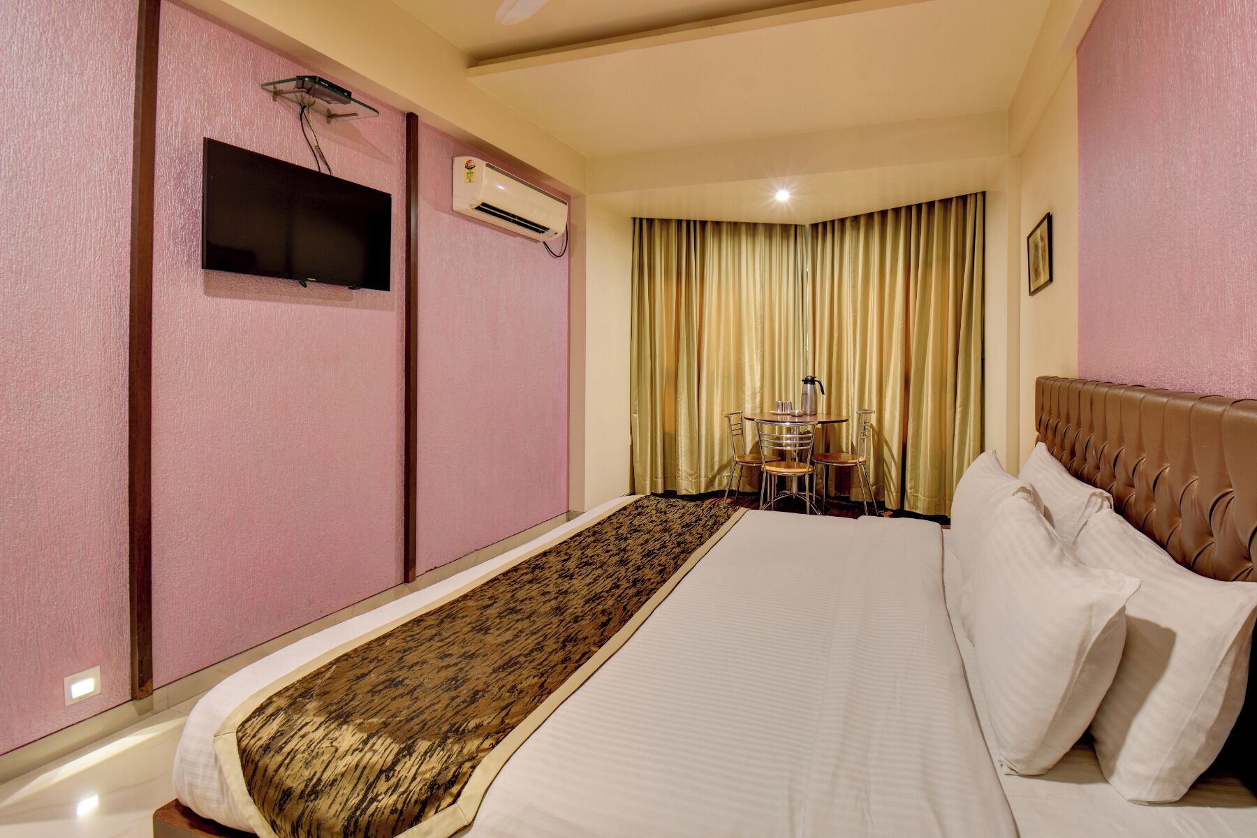 A hotel room with a bed, a small table with chairs, an air conditioning unit, and a television mounted on the wall.