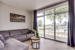 A modern living room with a grey sofa, a round wooden coffee table, and a large window with a view of a lake and houses. - Cube XL 77 | 6 persoons chalet in Zennewijnen (Zennewijnen)