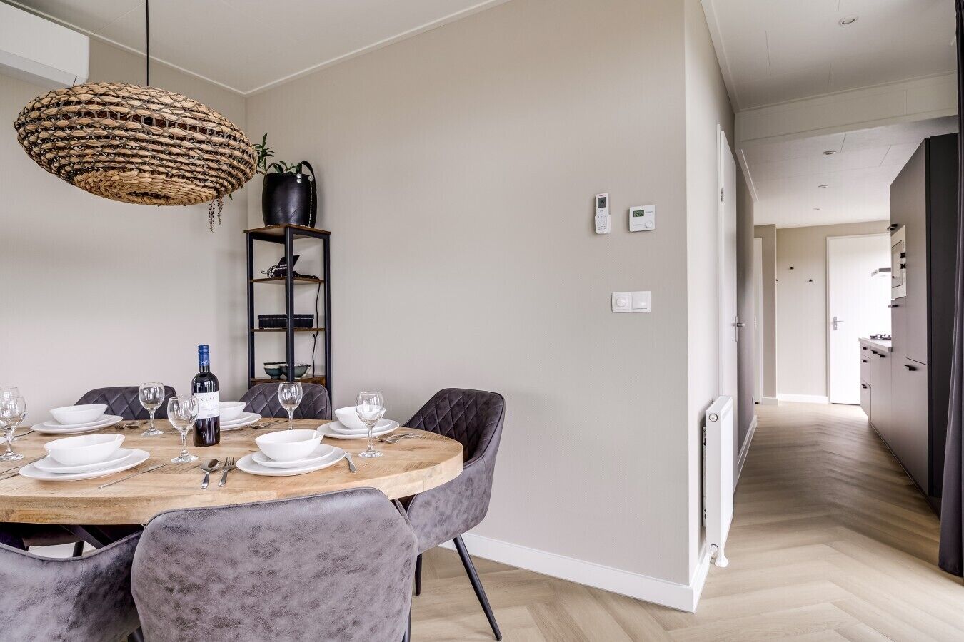 A dining area with a round wooden table, grey upholstered chairs, a woven pendant light, a black leather chair, and a black leather sofa.