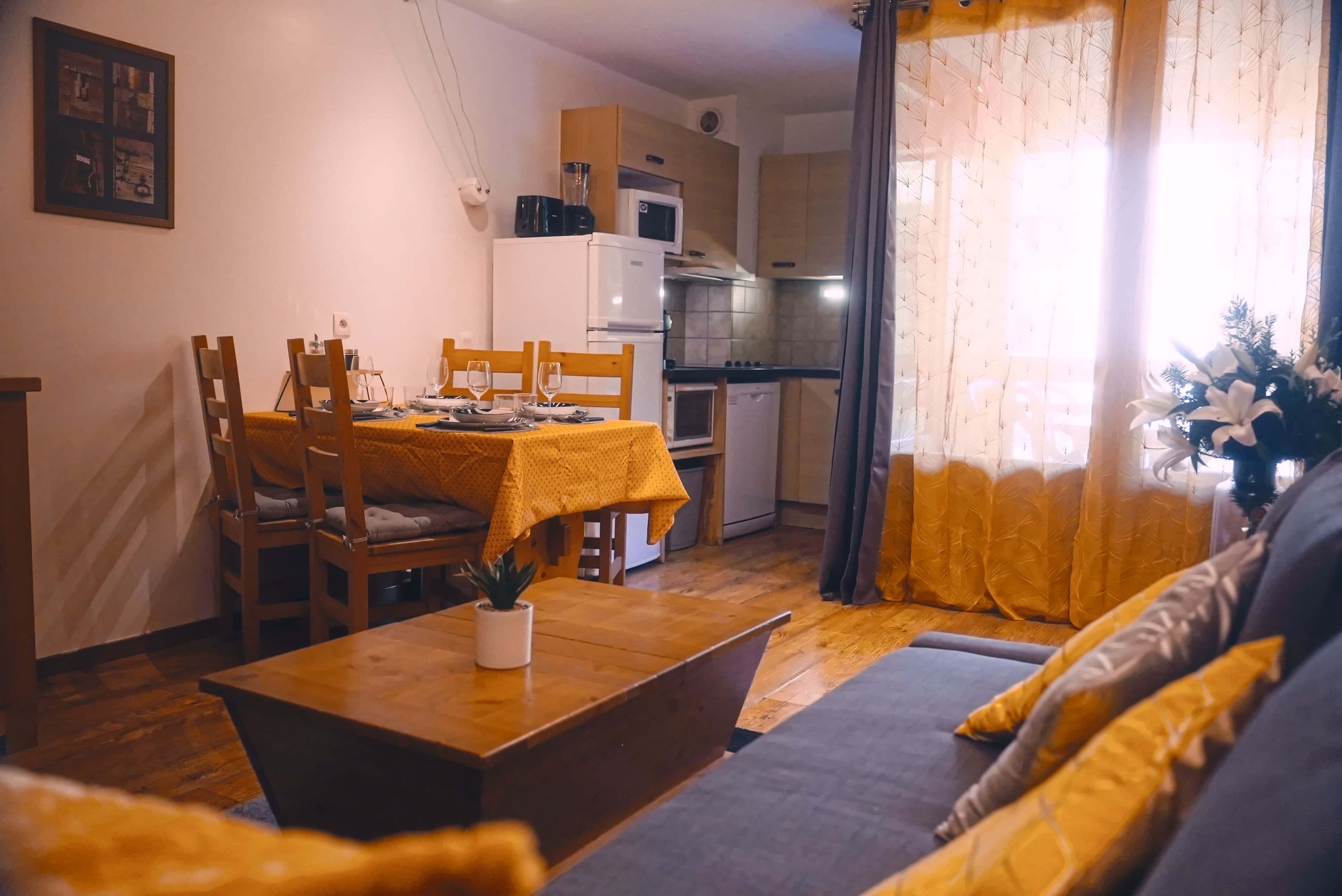 Ett vardagsrum med en soffa, ett matbord med stolar, ett kök i bakgrunden och ett fönster med gardiner.