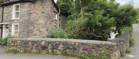 A stone building with a stone wall in front, a paved road, and trees in the background.