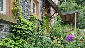Un edificio in pietra con un balcone in legno, rigogliosa vegetazione e un vaso di fiori.