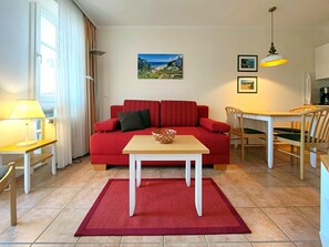 Un sofá rojo, una mesa de madera, una alfombra roja, una lámpara, una silla, una mesa de comedor y un cuadro en la pared.