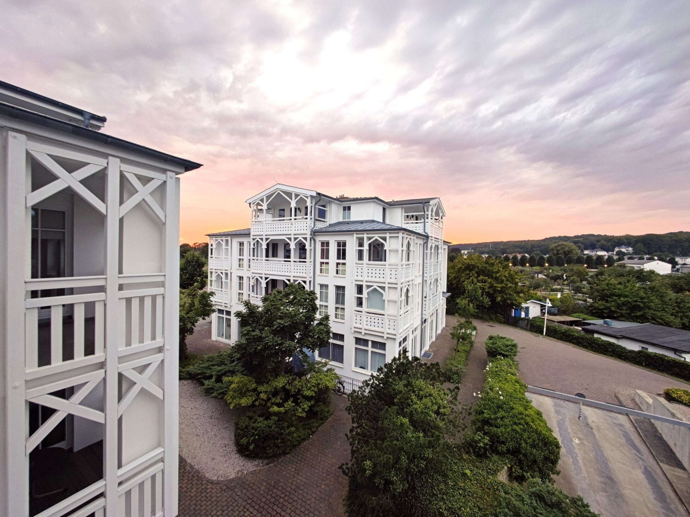 A white building with multiple balconies and a large windowed facade, surrounded by greenery and a paved driveway.