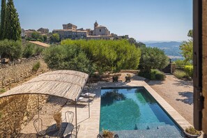 En pool med flisep terrasse, et overdækket siddeområde og udsigt til en historisk by i baggrunden.