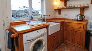Uma cozinha compacta com máquina de lavar, pia, armários de madeira, uma janela com vista e uma prateleira com utensílios de cozinha.