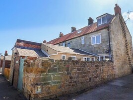 A stone building with a sloped roof, a small attached structure with a green door, and a stone wall with a sign.
