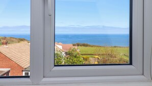Janela com vista para o mar, área gramada e um edifício com telhado vermelho.