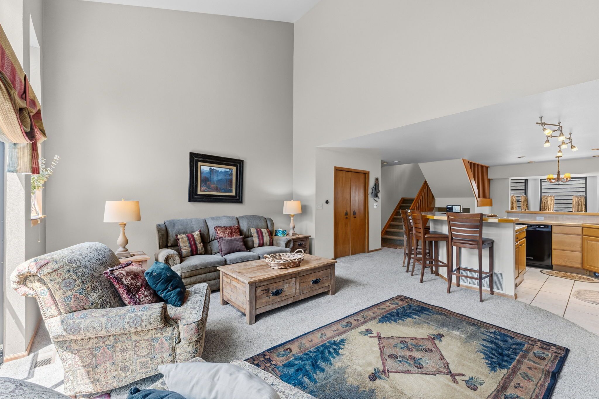 A spacious living area with a sofa, coffee table, and a rug. A kitchen with wooden cabinets and a dining area with bar stools is visible.