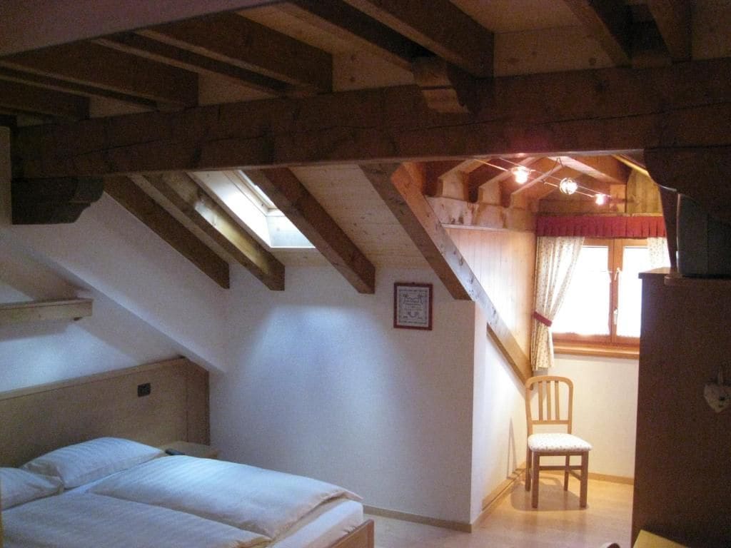 Une chambre avec un plafond en pente, un lit, une chaise et une fenêtre avec des rideaux.